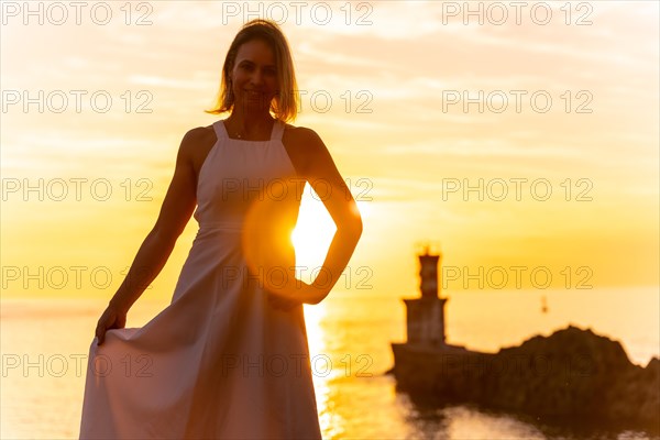 Portrait of a blonde woman smiling in a white dress at sunset next to a lighthouse in the sea