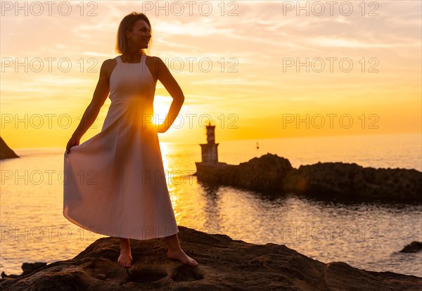 Portrait of a blonde woman in a white dress at sunset next to a lighthouse in the sea