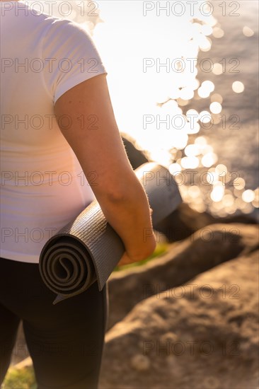 Hands of a woman with the yoga mat in nature by the sea at sunset