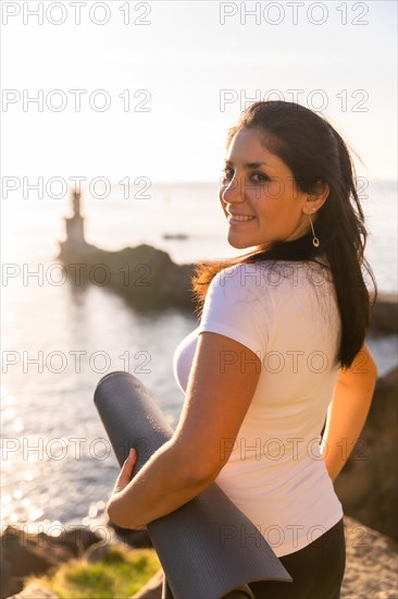 A woman with the yoga mat in nature by the sea at sunset