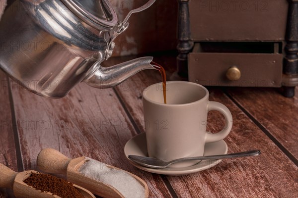 Metal vase pouring coffee into a ceramic cup on a wooden table