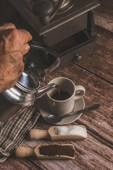 Metal vase pouring coffee into a ceramic cup on a wooden table