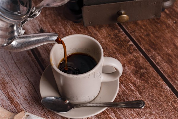 Metal vase pouring coffee into a ceramic cup on a wooden table