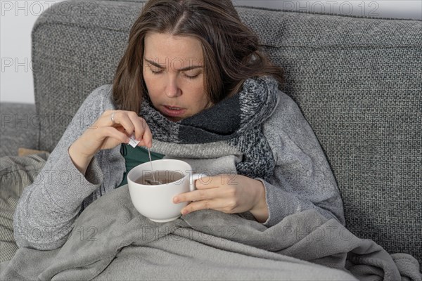 Sick woman covered with a blanket drinking an infusion on the couch in her home