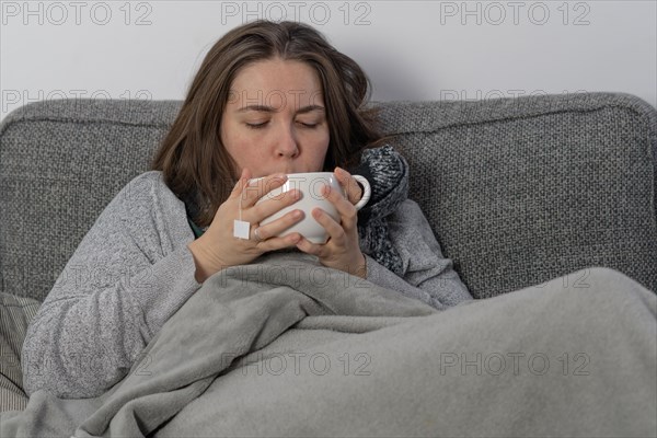 Sick woman covered with a blanket drinking an infusion on the couch in her home