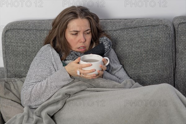 Sick woman covered with a blanket drinking an infusion on the couch in her home