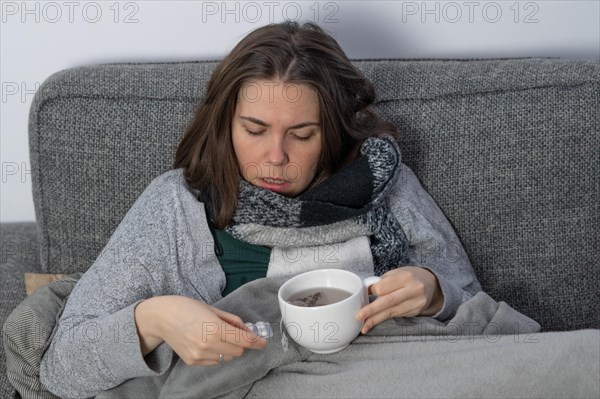 Sick woman covered with a blanket drinking an infusion on the couch in her home