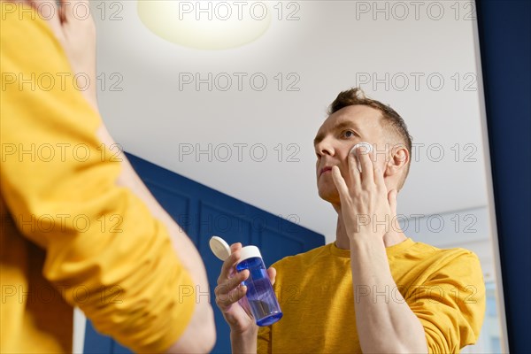 Middle aged man looking in the mirror and cleaning face with lotion using cotton pad