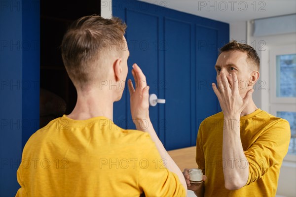 Middle-aged man applying anti-aging cream standing in bedroom in front of the mirror
