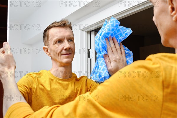 Positive middle-aged man cleaning mirror with cleaning cloth at home