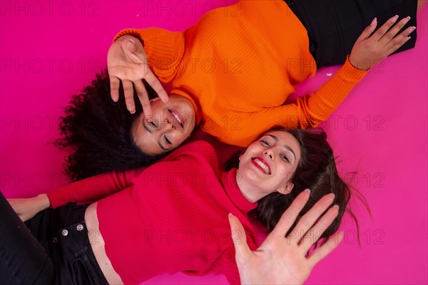 Two multi-ethnic women lying down smiling in a trendy posing on a pink background