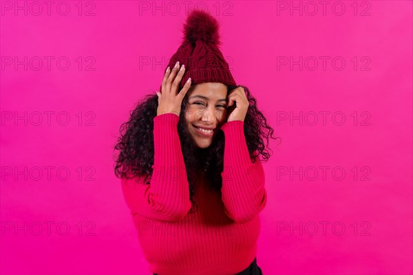 Curly-haired woman in a woolen cap on a pink background smiling shyly