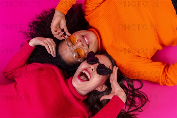 Two multi-ethnic women in sunglasses lying down smiling on a pink background