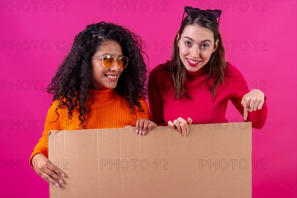 Two multiethnic female friends smiling and pointing at cardboard sign over pink background