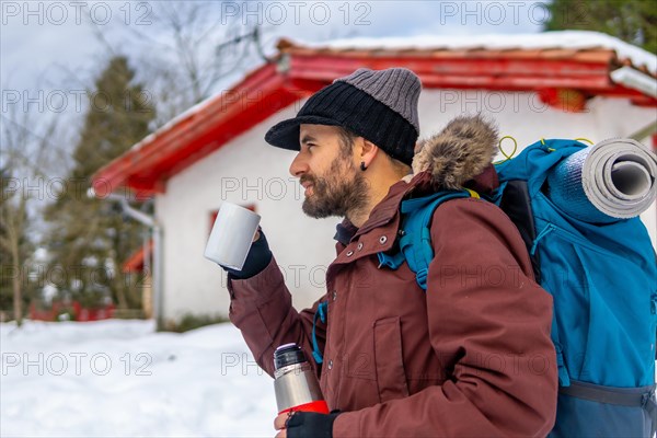 Man drinking coffee from a hot thermos in winter in the snow next to a cabin after sleeping in it