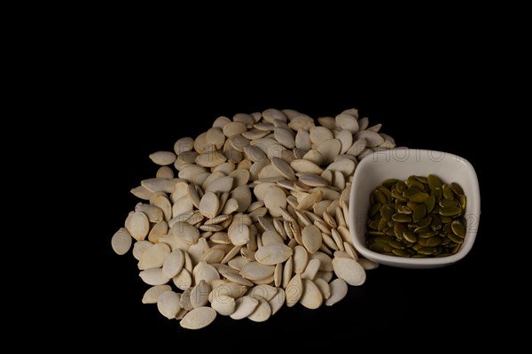 Roasted pumpkin seeds roasted with salt in a pile and seeds in a white bowl on a black background