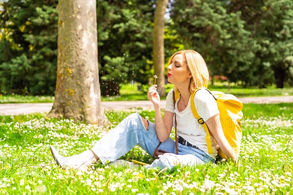 A blonde woman blowing on the dandelion plant in a park in the city