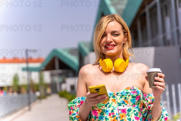 Portrait of a blonde woman walking with the phone through the city with yellow headphones and a take away coffee