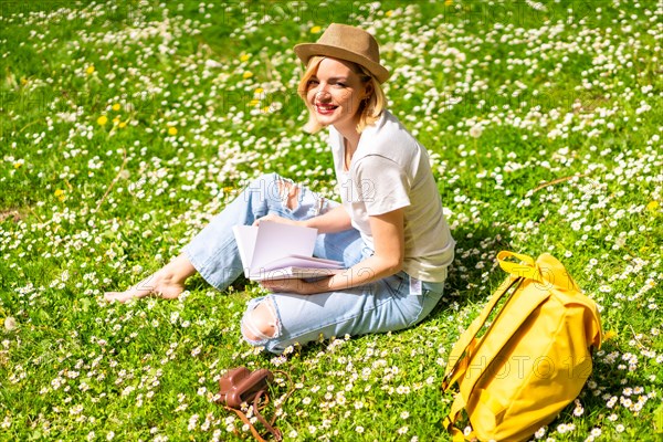 A young blonde girl in a hat writing poems in a notebook in spring in a park in the city