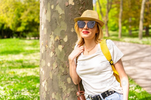 A tourist blonde woman with a hat and sunglasses leaning on a tree in the spring in a park in the city
