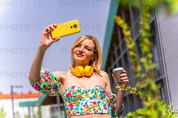 A blonde woman taking a selfie with her phone in the city with yellow headphones and a take away coffee