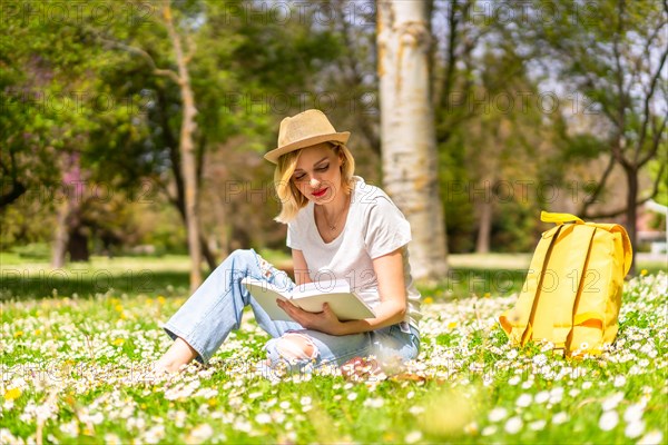 A young blonde girl in a hat reading a book in spring in a park in the city