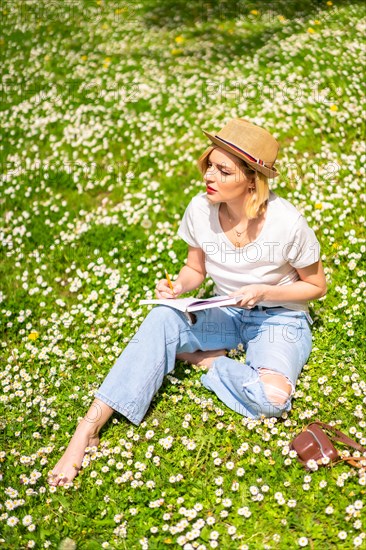 A young blonde girl in a hat writing poems in a notebook in spring in a park in the city