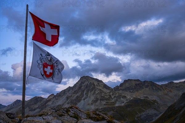 Swiss flag and flag of the SAC with cloudy sky at blue hour and Engadine mountains