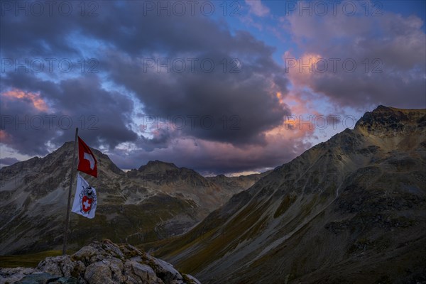 Swiss flag and flag of the SAC with cloudy sky at blue hour and Engadine mountains