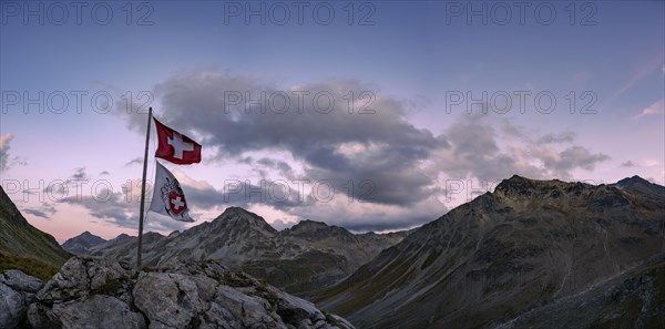 Swiss flag and flag of the SAC with cloudy sky at blue hour and Engadine mountains