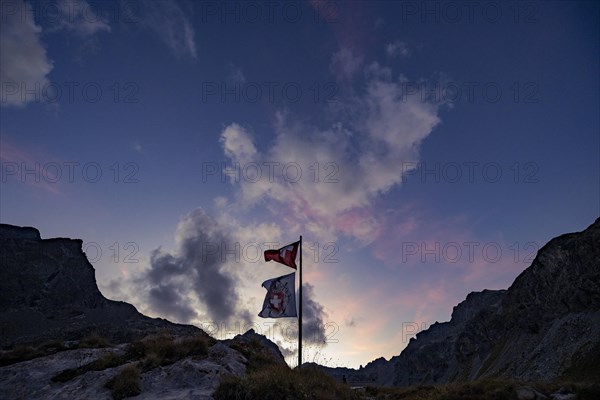 Swiss flag and flag of the SAC with cloudy sky at blue hour and Engadine mountains