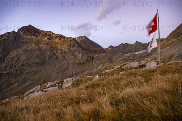 Swiss flag and flag of the SAC with cloudy sky at blue hour and Engadine mountains