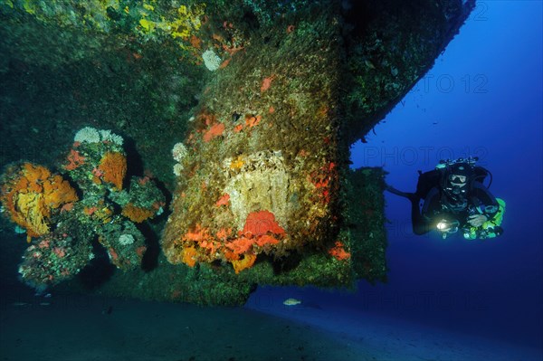 Diver with diving equipment for technical diving Tec Diving Tec Diving views illuminated looks at rudder propeller of sunken ship at bottom of Mediterranean Sea