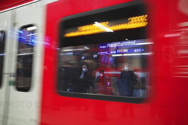 Travellers wearing Corona masks wait on the platform and are reflected in windows