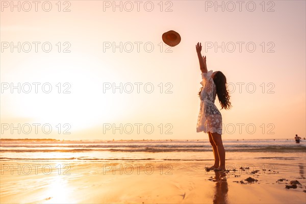 A woman in the sunset in a white dress at the sea in summer throwing the hat