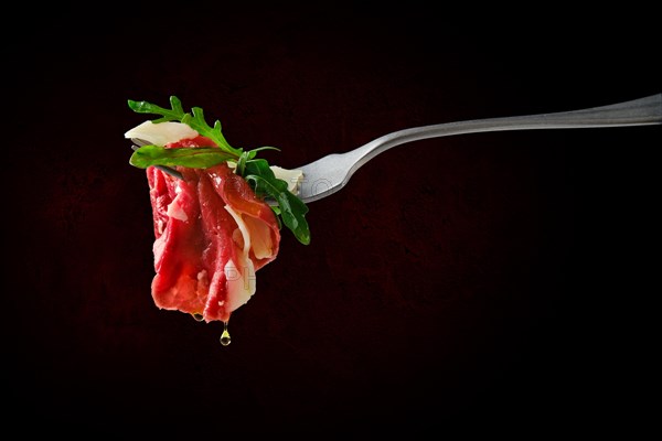 Piece of marbled beef carpaccio on fork with dripping olive oil over black background