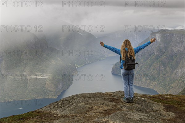 A girl stands on a mountain above the Aurlandsfjord in Norway and stretches her hands up in the air