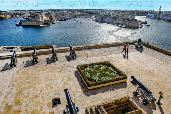 View over former parade ground with saluting cannons Battery Saluting Batter and flower bed in the form of Maltese Cross from historic Order of St. John next to Upper Barraka Garden on left Fort St. Angelo on Birgu Peninsula with village La Vittoriosa