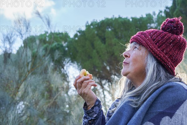 White-haired woman with red cap covered with a blanket eating an apple in the mountains