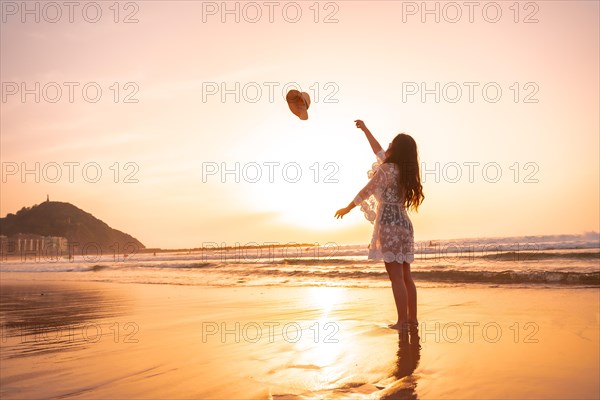 A woman in the sunset in a white dress at the sea in summer throwing the hat on vacation