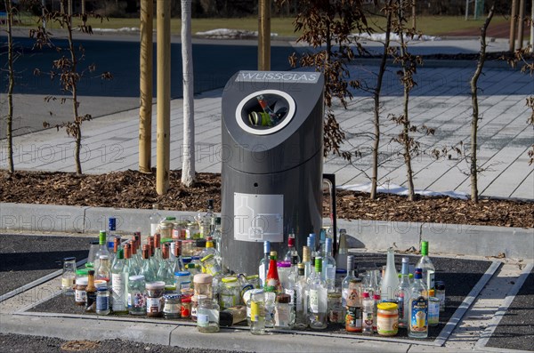 Bottles and jars stand in front of an overcrowded waste glass collection container