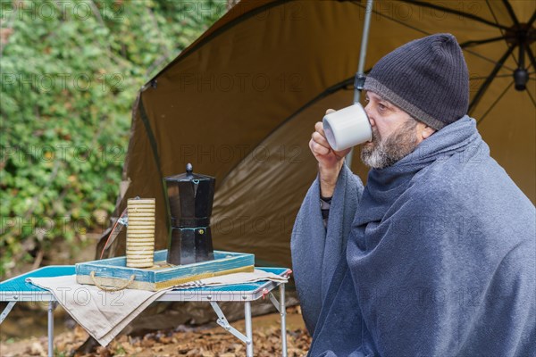 Bearded man covered with a camping blanket sipping a cup of coffee