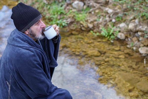 Bearded man covered with a blanket drinking coffee on the river bank