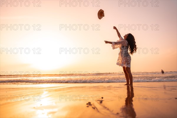 A woman in the sunset in a white dress at the sea in summer throwing the hat on vacation