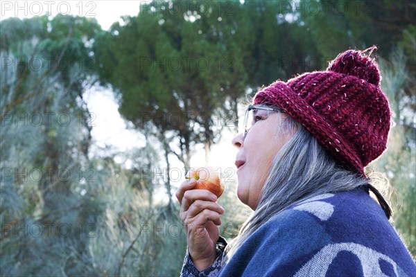 White-haired woman with red cap covered with a blanket eating an apple in the mountains