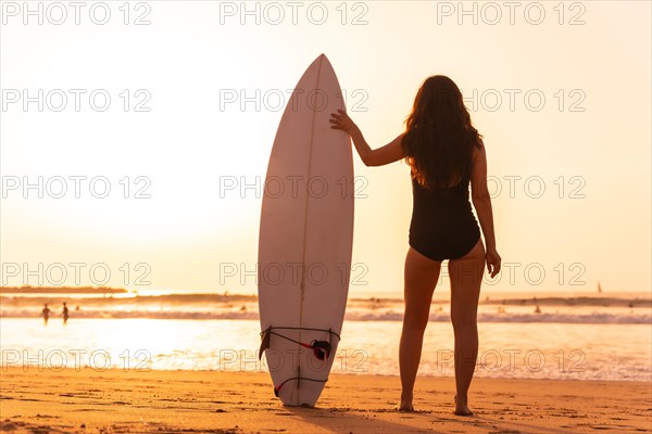 Surfer woman looking at the waves on a beach at sunset next to the surfboard
