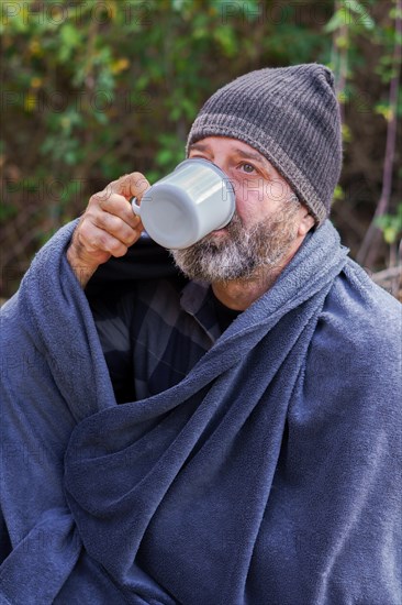 Bearded man covered with a camping blanket sipping a cup of coffee