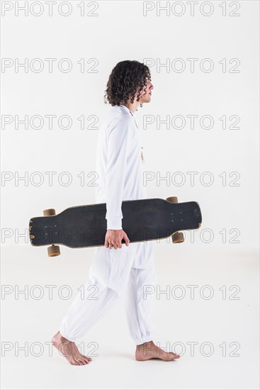 Side view of a young man with curly hair wearing white clothes and walking while holding a skate over white background