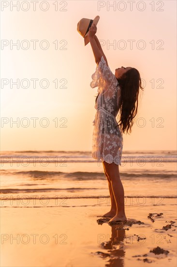 A woman in the sunset in a white dress at the sea in summer throwing the hat