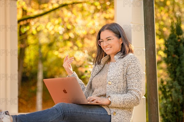 Pretty woman with glasses and MacBook in hand sits in the park at autumn time